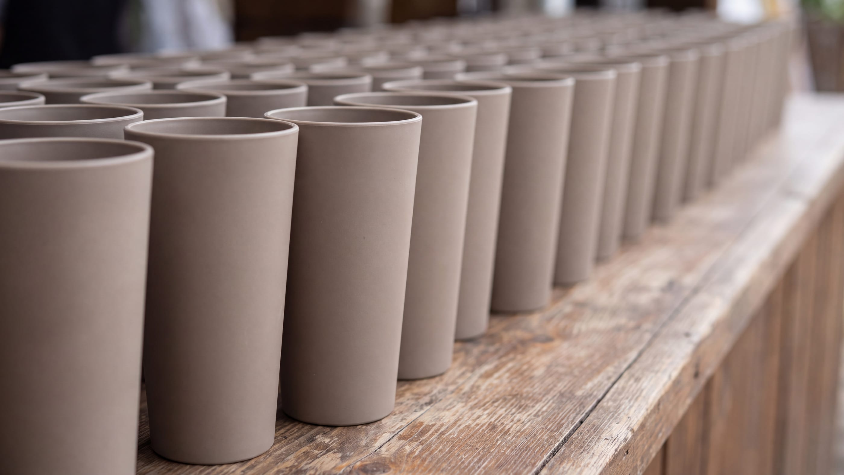 Lange Reihe gleicher unbedruckter Mehrwegbecher aus mattem Kunststoff dicht an dicht auf einer Holztheke im Freien, nach hinten unscharf werdend.
