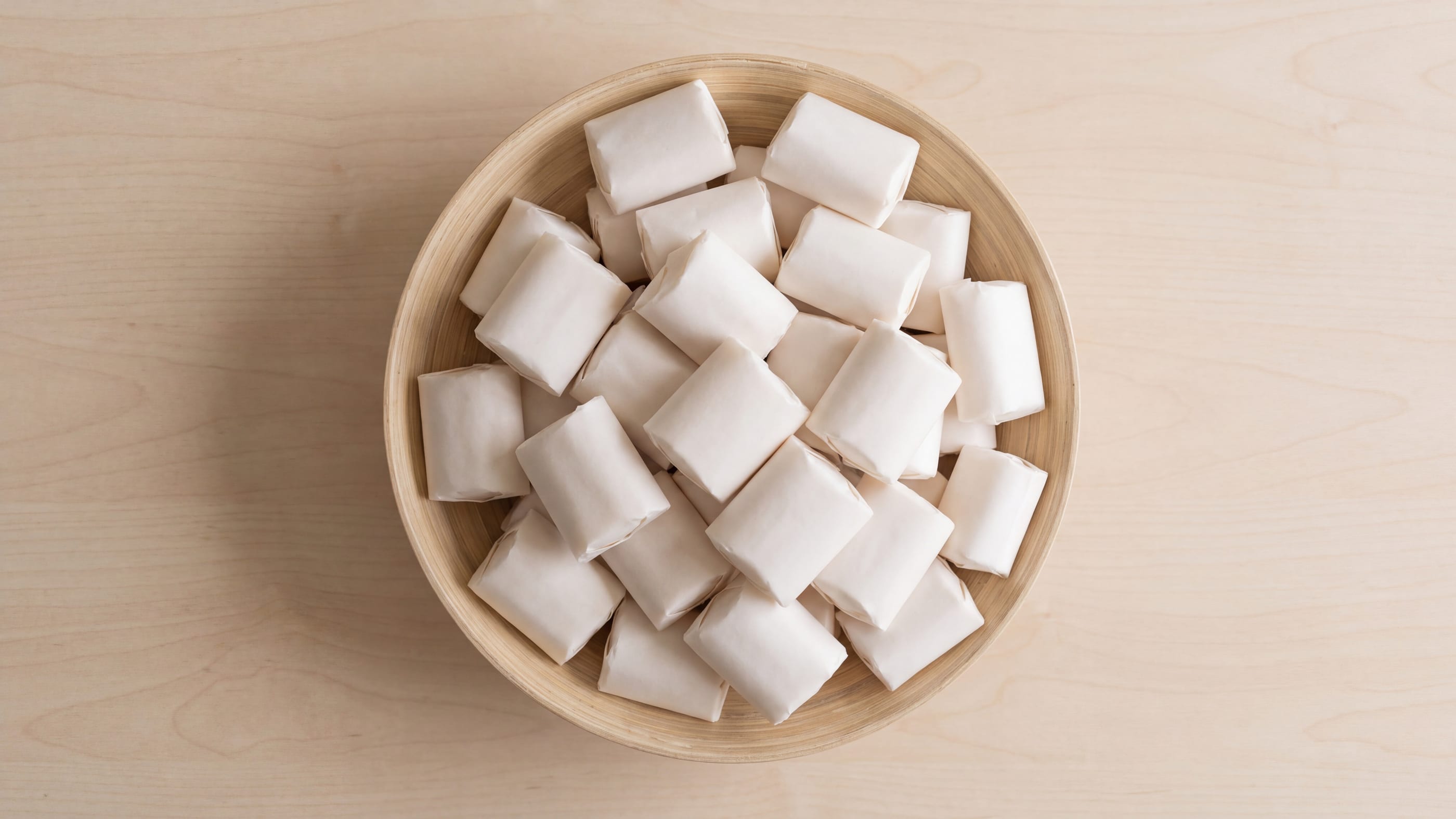 Flache Holzschale, dicht gefüllt mit einzeln in glattes unbedrucktes Papier eingeschlagenen kleinen Süßwaren, von oben gesehen.